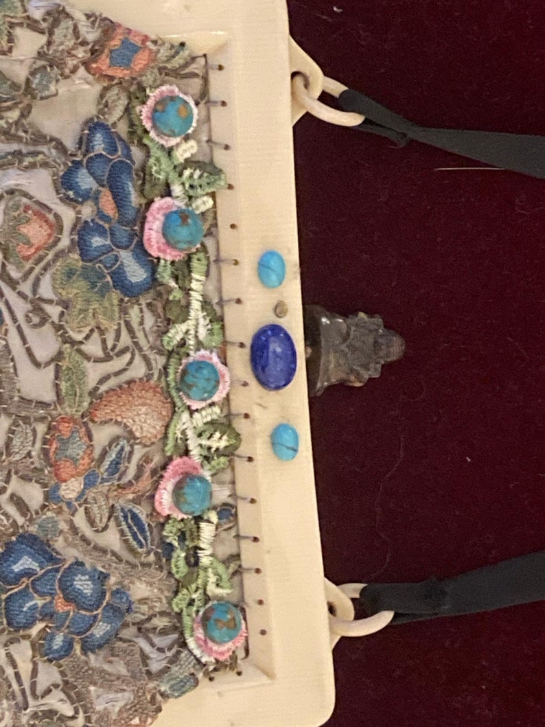 May include: Ivory-colored vintage handbag with intricate floral embroidery in blue, peach, and green. Embellished with turquoise and lapis lazuli stones. Features a cream-colored handle and black ribbon detail.