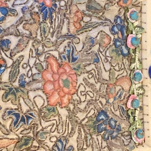 May include: Close-up of a section of embroidered fabric. The fabric features a complex floral design with blue, pink, and peach-colored flowers and green leaves on a cream background. The edges are trimmed with turquoise beads and pink flowers.