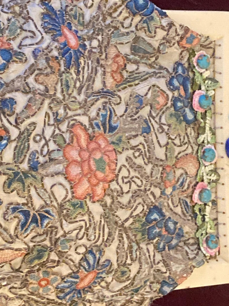 May include: Close-up of a section of embroidered fabric. The fabric features a complex floral design with blue, pink, and peach-colored flowers and green leaves on a cream background. The edges are trimmed with turquoise beads and pink flowers.