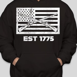 May include: Black hoodie featuring a distressed white graphic of the American flag with crossed rifles. The text "EST 1775" is below the flag. The hoodie has a front pocket and a hood.