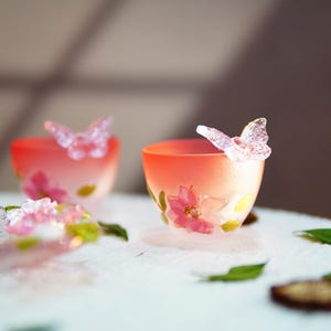 May include: Two small, decorative glass cups with a gradient orange and pink color scheme. Each cup features a clear, butterfly-shaped accent and floral designs. Green leaves and small brown pieces are scattered around the cups.