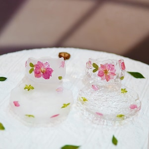 Peut inclure: Deux bagues en résine transparente avec des fleurs roses et des feuilles vertes incrustées, chacune posée sur un dessous de verre en résine transparente assorti. Sur une surface blanche, avec des pétales et des feuilles éparpillés.