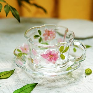 Op de afbeelding: Een heldere glazen theekop en schotel set, versierd met geperste roze bloemen en groene bladeren. De kop heeft een gebogen handvat en een geschulpte rand. De schotel weerspiegelt het bloemendesign van de kop.