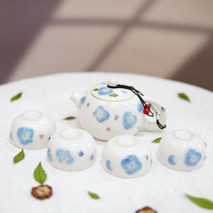 Op de afbeelding: Een witte keramische theepot en vier bijpassende kopjes, allemaal versierd met een delicaat blauw bloemmotief. De theepot heeft een zwart koordhandvat met rode kralen. De set is geplaatst op een wit oppervlak.