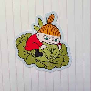 以下が含まれることがあります： オレンジ色の髪をまとめた赤色の衣装の漫画のキャラクターが、緑色の葉野菜の上に座っているステッカー。キャラクターは怒った表情をしています。ステッカーは水色の縁取りです。