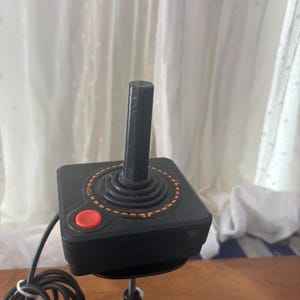 May include: A black video game joystick controller featuring a square base, a central vertical stick, and a single circular red button. An orange dashed circular graphic surrounds the base of the stick with the word TOP printed in orange text.