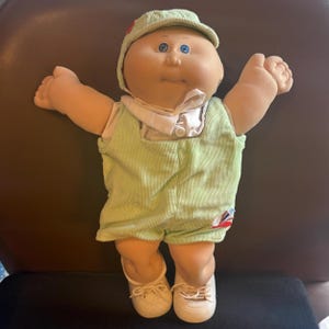 May include: A Cabbage Patch Kid doll wearing a light green corduroy outfit, a matching cap, and white shoes. The doll has blue eyes and a smiling expression, with arms outstretched. A classic collectible toy.