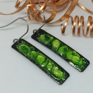 May include: A pair of handmade earrings with a rectangular shape. The earrings feature a dark border with a vibrant green, textured design. The ear wires are silver-colored. Decorative copper-colored metal curls are in the background.