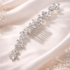 Può includere: Un pettine per capelli da sposa argentato con un design floreale. Il pettine presenta cristalli trasparenti e scintillanti disposti a forma di foglia. Il pettine è posto su uno sfondo di tessuto di raso bianco.