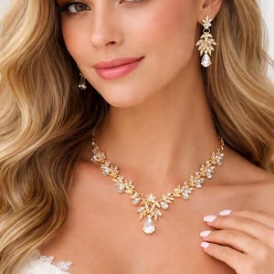 Può includere: Una collana dorata e orecchini abbinati. I gioielli presentano un design floreale con cristalli chiari e scintillanti. La collana ha un pendente a forma di goccia. La donna indossa un abito bianco.
