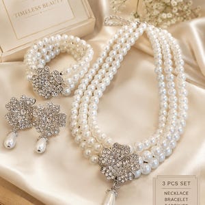 Könnte beinhalten: Ein dreiteiliges Schmuckset bestehend aus einer mehrreihigen Perlenkette, einem mehrreihigen Perlenarmband und zwei Ohrhängern. Jedes Schmuckstück verfügt über einen silberfarbenen, blumenförmigen Verschluss mit klaren Kristallen.