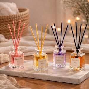 Op de afbeelding: Vier glazen rietverspreiders met verschillende gekleurde rietjes en vloeistof. De verspreiders zijn in tinten roze, geel, paars en oranje. Elke verspreider heeft een label met de tekst "FAGOTYDE". De verspreiders staan op een marmeren oppervlak.