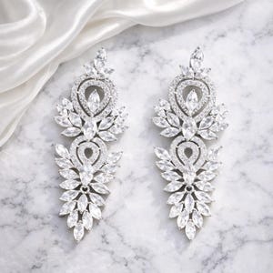 Pode incluir: Um par de brincos pendurados prateados com um design floral. Cada brinco apresenta um topo em forma de coração, uma pedra oval central e múltiplas pedras claras e brilhantes dispostas em um padrão de folhas. Os brincos são exibidos em uma superfície de mármore.
