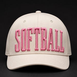 Op de afbeelding: Een beige honkbalpet met het woord SOFTBALL in reliëf geborduurd in roze letters op de voorpanelen. De pet heeft een gestructureerde kroon, een gebogen klep en kleine ventilatiegaatjes op de bovenste panelen van de pet.