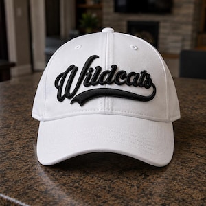 May include: A white baseball cap featuring the word Wildcats in a raised black script font across the front panels. The cap has a structured crown, a curved brim, and a button at the top center, constructed from a solid white fabric material.