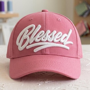 Puede incluir: Una gorra de béisbol de color rosa empolvado con la palabra Blessed bordada en letras cursivas blancas en relieve en los paneles frontales. La gorra tiene una corona estructurada, una visera curva y costuras a juego en toda la tela.