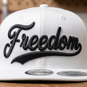 May include: A white baseball cap featuring the word Freedom embroidered in black script across the front panel. The cap has a flat brim with two circular stickers attached to the top, and the fabric has a visible woven texture throughout the crown.