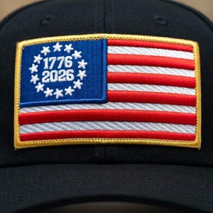Peut inclure: Casquette de baseball noire avec un écusson représentant un drapeau américain stylisé. L'écusson du drapeau comprend des rayures rouges et blanches, un champ bleu avec des étoiles blanches et les chiffres "1776 2026". L'écusson est bordé d'or.