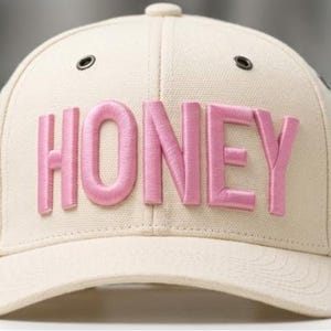 Może przedstawiać: Beżowa czapka bejsbolowa z napisem "HONEY" wyszytym na różowo z przodu. Czapka ma zakrzywiony daszek, guzik na górze i dwa małe otwory wentylacyjne. Kapelusz jest wykonany z teksturowanej tkaniny.