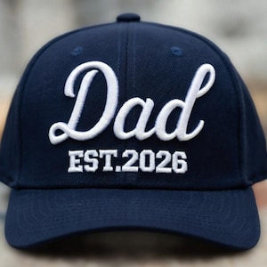 May include: A navy blue baseball cap with the word "Dad" embroidered in white script. Below the word "Dad" is the text "EST.2026" in white block letters. The cap has a curved brim and a button on top.
