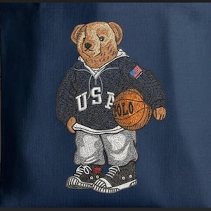 Op de afbeelding: Geborduurd ontwerp van een beer met een marineblauwe hoodie met de tekst "USA POLO" en een Amerikaanse vlagpatch. De beer houdt een basketbal vast en draagt grijze joggingbroek en zwarte sneakers. De achtergrond is effen marineblauw.