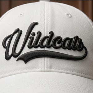 May include: White baseball cap with the word "Wildcats" embroidered in black script across the front. The hat has a curved brim and a structured crown. A second baseball cap is visible in the background.