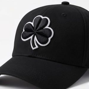 Puede incluir: Gorra de béisbol negra con un emblema de trébol negro con contorno blanco en la parte delantera. La gorra tiene una visera curva y un botón en la parte superior. El trébol, un trébol de tres hojas, es un símbolo de suerte y herencia irlandesa.