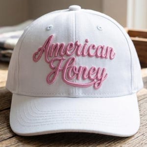 Puede incluir: Una gorra de béisbol blanca con las palabras American Honey bordadas en letras cursivas rosas en relieve en el panel frontal. La gorra tiene una corona estructurada, una visera curva y un pequeño botón blanco en la parte superior.