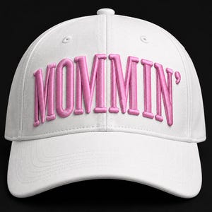 May include: A white baseball cap featuring the word MOMMIN' embroidered in raised, bright pink lettering across the front panels. The cap has a structured crown, a curved brim, and a matching white button at the top center of the headpiece.