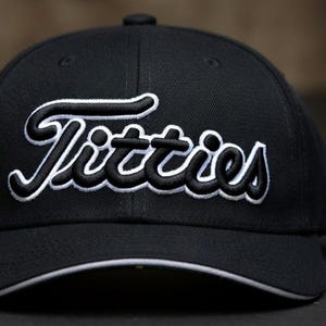 May include: Black baseball cap with the word "Titties" embroidered in white and black script across the front. The cap has a curved brim with a white trim and a black button on top.