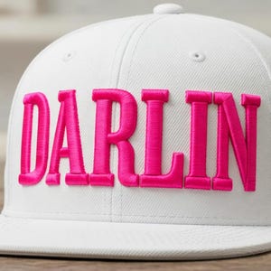 Op de afbeelding: Witte baseballcap met het woord "DARLIN" geborduurd in felroze. De cap heeft een platte klep en een ronde kroon. Een tweede witte cap is gedeeltelijk zichtbaar op de achtergrond.