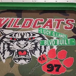 May include: Camouflage fabric with a green border featuring the word "WILDCATS" in red glitter lettering. A tiger face with red eyes and the text "LUCY C. LANEY BLVD BUILT" is embroidered. A red paw print with the number "97" is also visible.