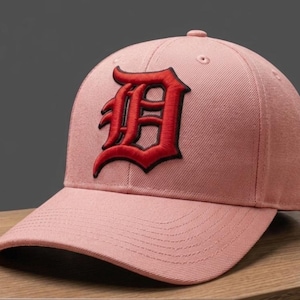 May include: A pink baseball cap with a curved peak. The cap has a red embroidered letter "D" on the front. The hat is made of a textured fabric and has a button on top.