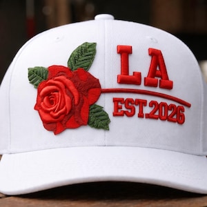 Puede incluir: Gorra de béisbol blanca con una rosa roja y hojas verdes bordadas en la parte delantera. La gorra presenta el texto rojo "LA EST 2026". La gorra tiene una visera curva y un botón en la parte superior.