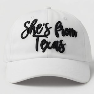 May include: White baseball cap with the text "She's from Texas" in black script. The hat has a curved brim and a button on top. A stylish accessory for anyone from Texas.