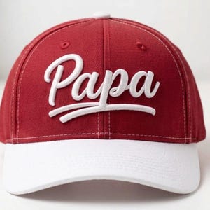 May include: A red baseball cap with a white brim. The word "Papa" is embroidered in white script across the front. The hat has a curved brim and a structured crown, suitable for casual wear.