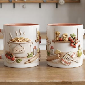 Könnte beinhalten: Zwei weiße Keramikbecher mit Pasta-Motiv. Jeder Becher hat einen pfirsichfarbenen Griff und Innenraum. Ein Becher zeigt einen Topf Spaghetti mit dem Namen "Max". Der andere Becher hat Nudeln, Tomaten und ein Regal mit Küchenutensilien.