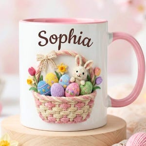 Puede incluir: Taza de cerámica blanca con interior y asa rosa, con un diseño de cesta de Pascua. La cesta contiene huevos coloridos, un conejito y flores. El nombre "Sophia" está impreso en la taza.