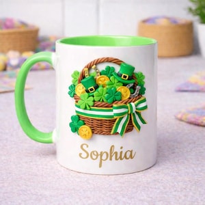 Puede incluir: Taza de cerámica blanca con interior y asa verdes. La taza presenta un diseño del Día de San Patricio con una cesta de tréboles, monedas de oro y sombreros de duende. El nombre "Sophia" está impreso en letras doradas.