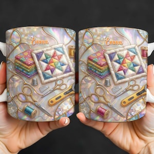 K&ouml;nnte beinhalten: Zwei wei&szlig;e Keramikbecher mit Quilt-Motiv. Jeder Becher zeigt einen bunten Quiltblock, Stoffstapel, Scheren und Garnrollen. Der Name "Emma" ist in Schreibschrift auf jedem Becher geschrieben.