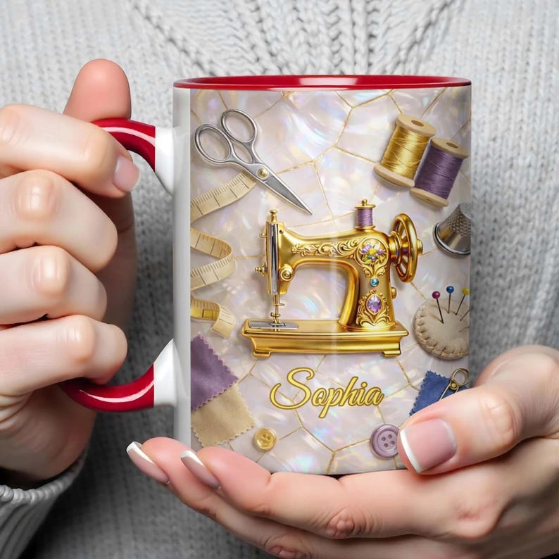 K&ouml;nnte beinhalten: Eine wei&szlig;e Keramik-Tasse mit rotem Innenraum und Henkel, verziert mit einer goldenen N&auml;hmaschine, Schere, Garnrollen und dem Namen "Sophia". Die Tasse hat einen perlmuttartigen Hintergrund und wird von einer Person gehalten.