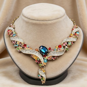 Pode incluir: Um colar dourado com uma grande pedra preciosa azul oval no centro. O colar apresenta uma cascata de pedras preciosas coloridas e cristais transparentes, criando um design brilhante e ornamentado. O colar é exibido em um manequim.