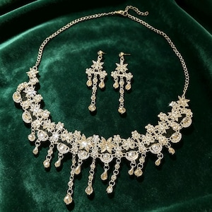 May include: A silver-toned necklace and matching pair of drop earrings featuring intricate floral and star-shaped metalwork encrusted with clear crystals. The necklace includes dangling crystal accents and a delicate chain with an adjustable clasp.