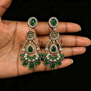 May include: A pair of gold-toned chandelier earrings with green and clear gemstones. The earrings feature oval, teardrop, and round-cut green stones, accented with clear crystals. The design includes floral and leaf-like elements.