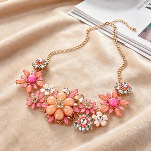 Puede incluir: Collar de cadena dorada con diseño floral. El collar presenta un grupo de colgantes de flores en tonos rosa, melocotón y blanco, con detalles de pedrería. Un accesorio llamativo.