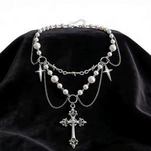 Peut inclure: Un collier argenté orné de fausses perles, de chaînes et de breloques en forme d'étoiles. Le bijou comprend un pendentif en forme de croix et un élément en fil de fer barbelé. Présenté sur un fond noir.