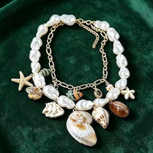May include: A layered necklace featuring large, irregular white pearls and gold-colored chain. The necklace is adorned with various seashells, a starfish, and other ocean-themed charms. The background is a dark green velvet.