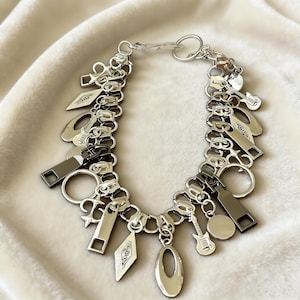 May include: Silver-toned necklace with a zipper-link chain design. The necklace features various charms, including guitars, zippers, and geometric shapes. The necklace is displayed on a soft, off-white fabric.