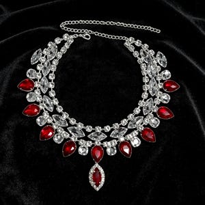 May include: A multi-layered silver-tone necklace featuring clear marquise-cut rhinestones and teardrop-shaped red crystals. The design includes a central dangling red crystal pendant and an adjustable chain with a lobster claw clasp closure.