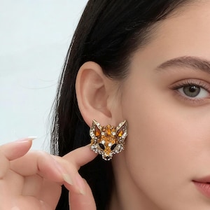 Może przedstawiać: Złote kolczyki w kształcie lisa. Kolczyki mają bursztynowe i przezroczyste kryształowe akcenty z czarnymi kryształowymi oczami. Twarz lisa jest szczegółowa z misternym rozmieszczeniem kryształów, tworząc efekt iskrzenia.